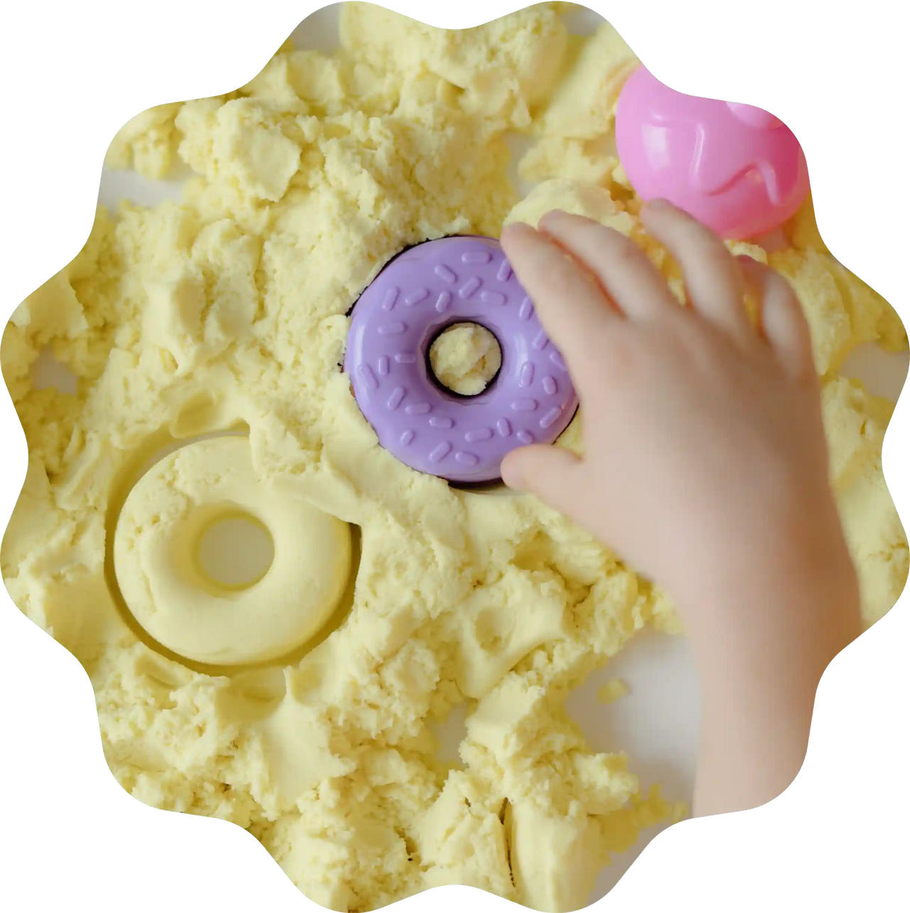 Kinderhand spielt mit Donut-Formen aus gelbem Sandolin Zaubersand, violette und rosa Förmchen liegen auf dem Tisch.