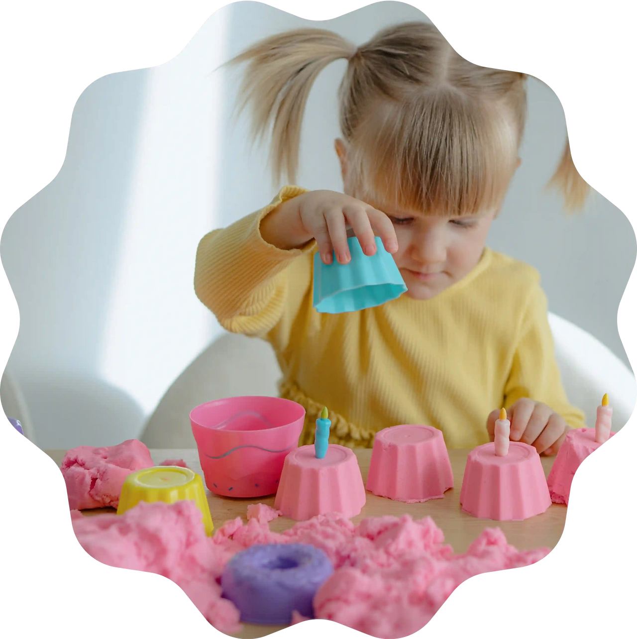 Mädchen formt bunte Cupcakes aus rosa Sandolin Zaubersand mit Förmchen und Kerzen am Tisch.